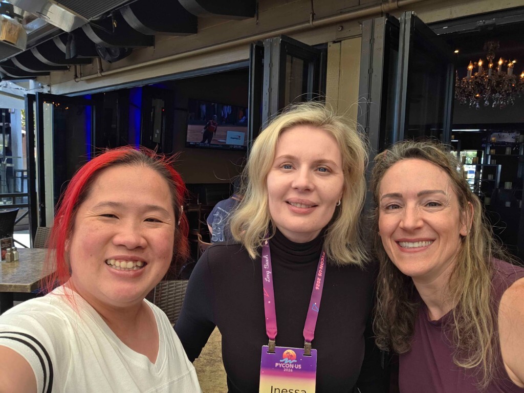 Three pyOpenSci community members smiling together at an outdoor patio during PyCon US 2026 in Long Beach. The person in the center wears a PyCon US 2026 conference lanyard and name badge.