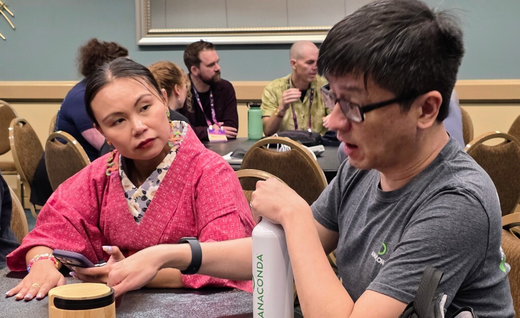 Two attendees have a focused one-on-one discussion at a table during the PyCon US Maintainer Summit, leaning in as they talk through a shared challenge.