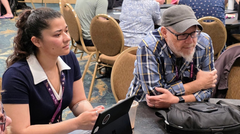 Two maintainers sit side by side at a round table during the PyCon US Maintainer Summit, focused on a session with laptops and conference lanyards on the table.